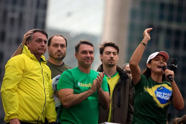 Michelle Bolsonaro, esposa del expresidente Jair Bolsonaro, habla mientras sobre un vehículo en un mitin, mientras el candidato presidencial, Flávio Bolsonaro, aplaude y el hermano de este, Carlos, observa.